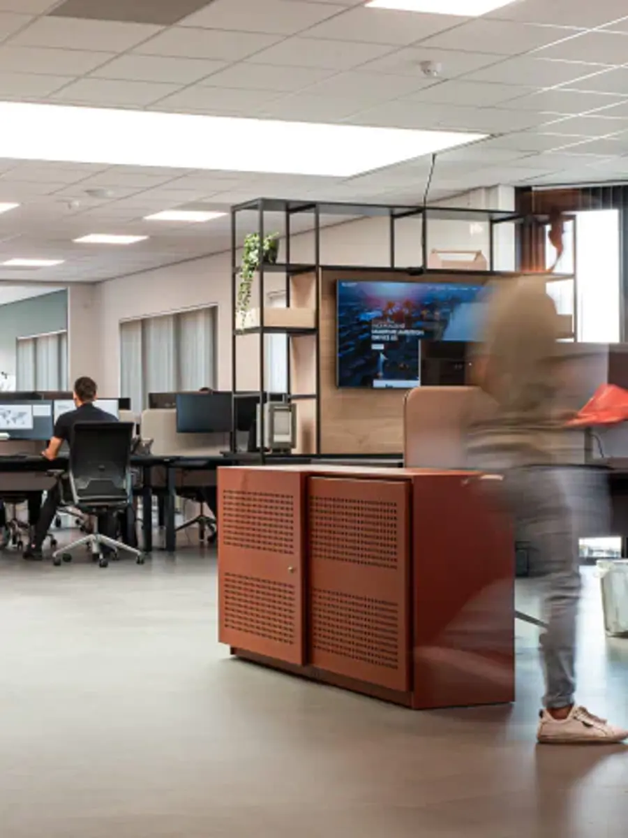 A woman in a white shirt is walking in a room with a brown desk.