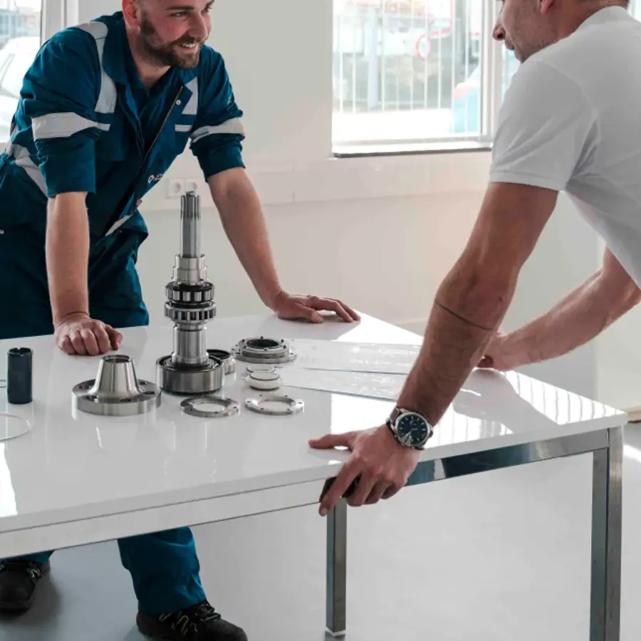 Two men in blue overalls are standing in front of a table with a motorcycle engine on it.